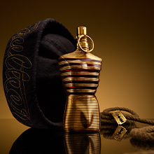 Jean Paul Gaultier Le Male Elixir in goldener Flasche, dargestellt mit schwarzer Matrosenmütze und goldenem Seil auf reflektierender Oberfläche.