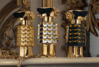 Drei goldene Parfumflakons in Form stilisierter Roboterfiguren stehen nebeneinander auf einem verzierten Kaminsims. Ihre Körper sind mit glänzenden Schuppenmustern in Gold, Weiß und Dunkelblau verziert, die Köpfe tragen schwarze Sonnenbrillen. Im Hintergrund opulente, goldene Dekoration.