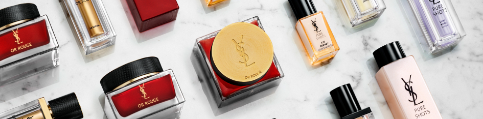 Kategoriebanner Pflege von Yves Saint Laurent: Verschiedene YSL-Pflegeprodukte wie Cremetiegel, Flakons und Pflegeflaschen in Rot-, Gold- und Beigetönen liegen auf einer hellen Marmoroberfläche.